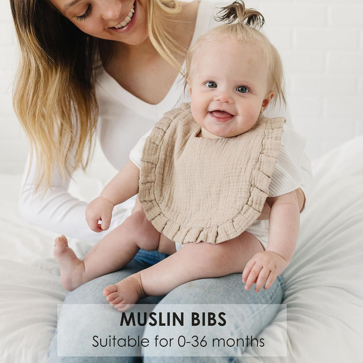 Smiling baby wearing a beige muslin bib with ruffled edges, sitting on an adult's lap on a white surface. Text overlay reads 'MUSLIN BIBS - Suitable for 0-36 months.'