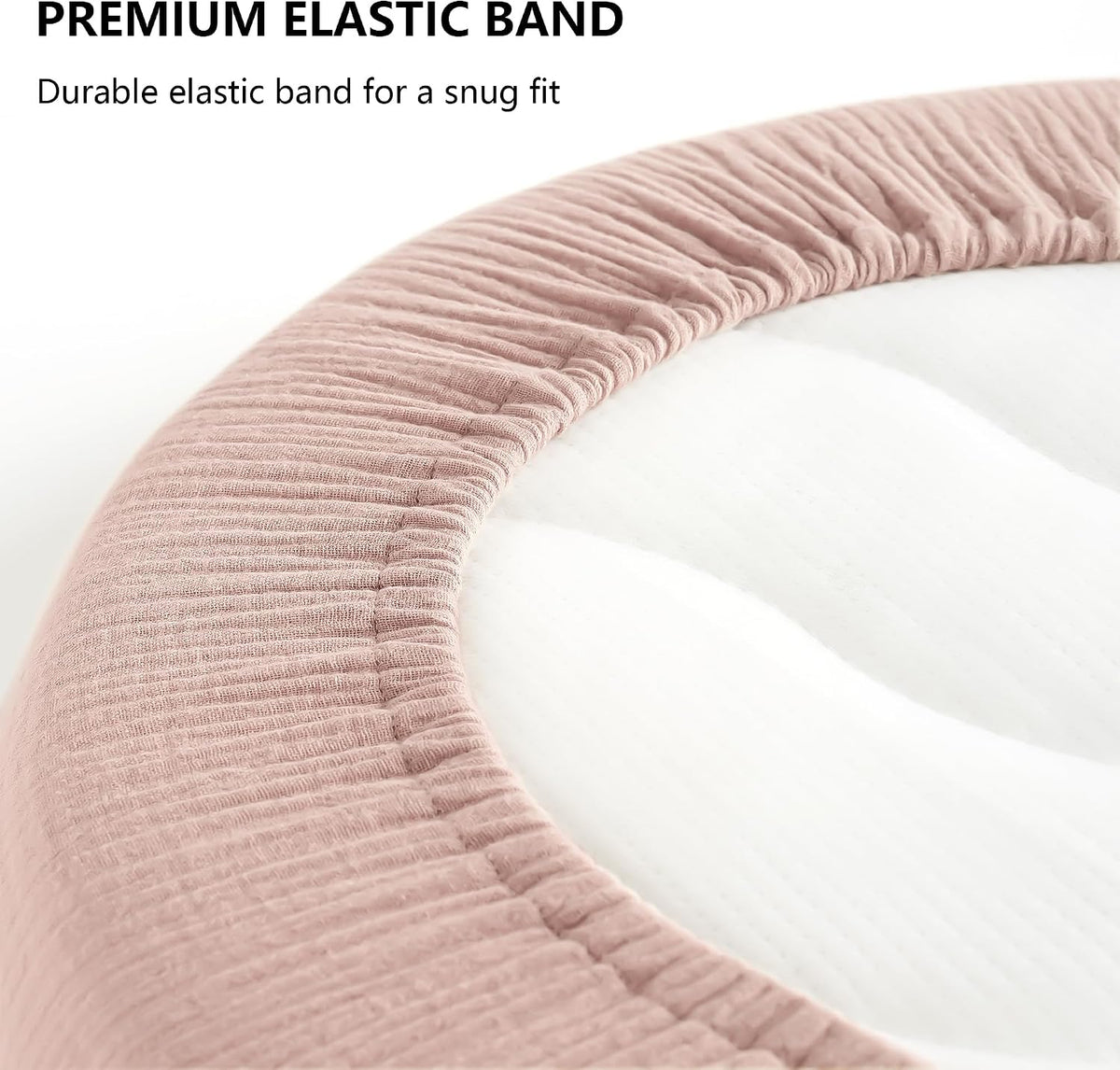 Close-up of a dusty pink fitted bassinet sheet showing ribbed muslin texture and elastic band edge wrapping around a white oval mattress. Text reads 'PREMIUM ELASTIC BAND - Durable elastic band for a snug fit.'
