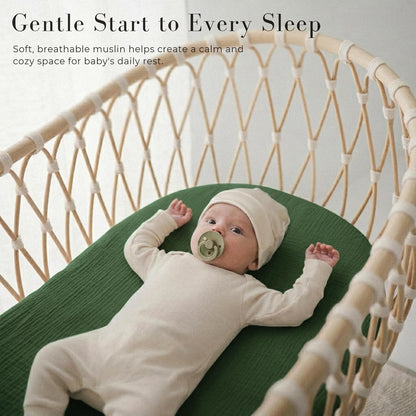 Baby lying in a wicker bassinet on a forest green fitted muslin sheet, wearing a cream outfit and hat with a pacifier. Text reads 'Gentle Start to Every Sleep' with a breathable muslin tagline.