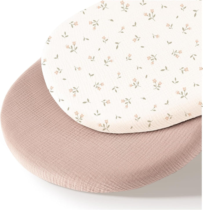 Two oval bassinet fitted sheets stacked together. The top sheet is ivory with a small scattered floral print in pink and sage green. The bottom sheet is solid dusty rose pink with a visible muslin texture.