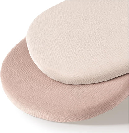 Two oval fitted bassinet sheets in a textured muslin fabric, stacked slightly offset. The top sheet is ivory/cream and the bottom is dusty pink, both showing a fine woven crinkle texture.