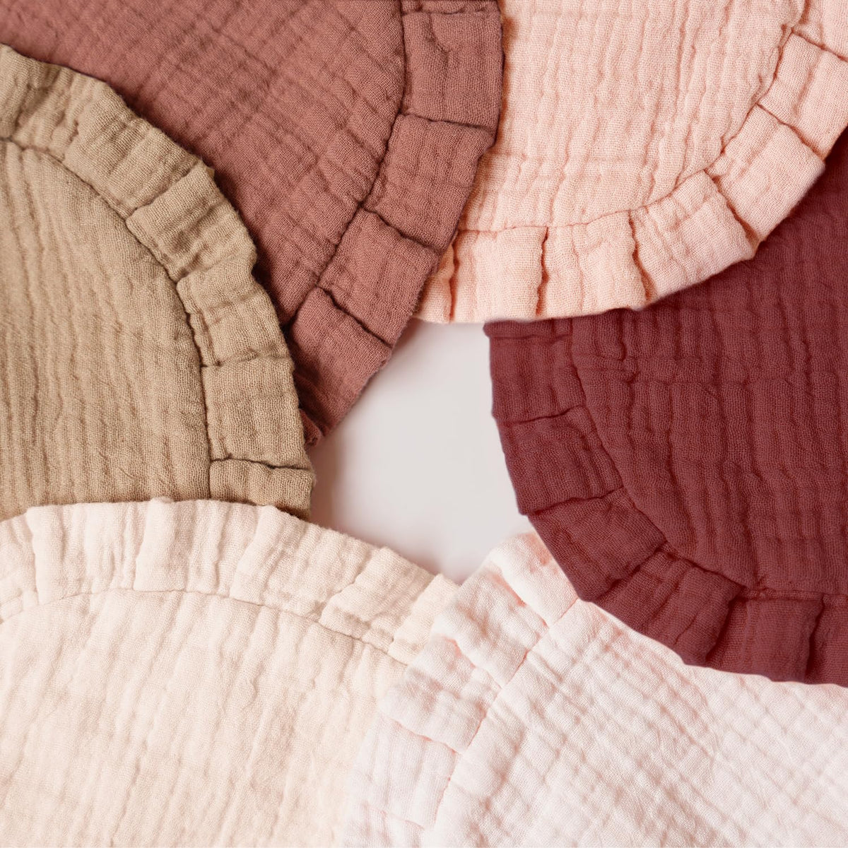 Five muslin cotton baby bibs with ruffled edges arranged in a fan pattern on a white surface, shown in five colors: tan, mauve, light pink, dark red, and pale blush.