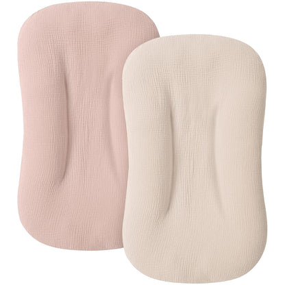 Two baby lounger covers in muslin fabric shown side by side: a dusty pink cover on the left and a cream/ivory cover on the right. Both have a rounded rectangular shape with a textured woven surface.