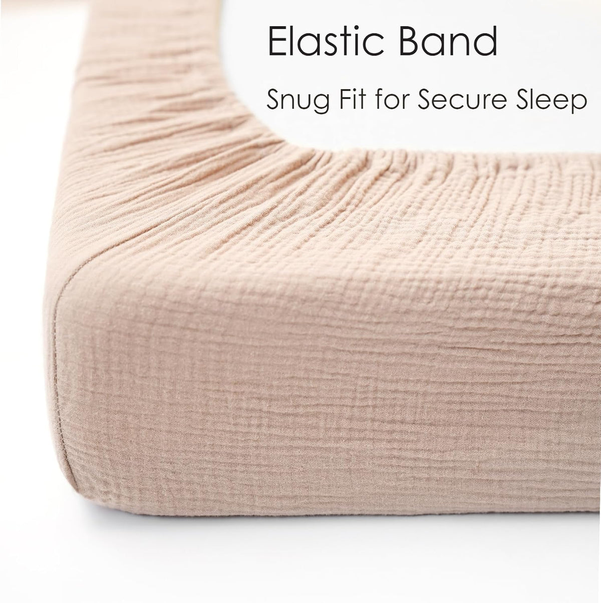 Close-up corner view of a fitted crib sheet in soft blush pink muslin fabric, showing gathered elastic band along the top edge. Text reads 'Elastic Band - Snug Fit for Secure Sleep.'