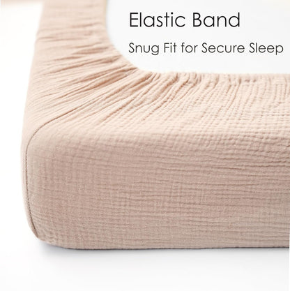 Close-up corner view of a fitted crib sheet in soft blush pink muslin fabric, showing gathered elastic band along the top edge. Text reads 'Elastic Band - Snug Fit for Secure Sleep.'