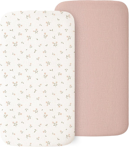 Two muslin fitted bassinet sheets with rounded corners. The front sheet is white with a scattered small pink floral and green leaf pattern. Behind it is a solid dusty pink sheet with a subtle textured weave. Both have a soft, woven fabric appearance.