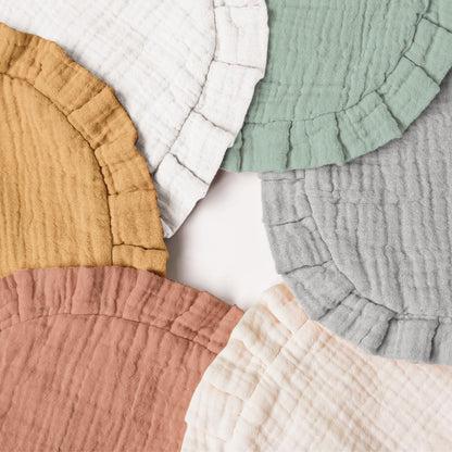 Five round muslin cotton bibs with ruffled edges arranged in a fan-like spread, shown in mustard yellow, white, sage green, light gray, dusty rose, and cream colorways on a white background.