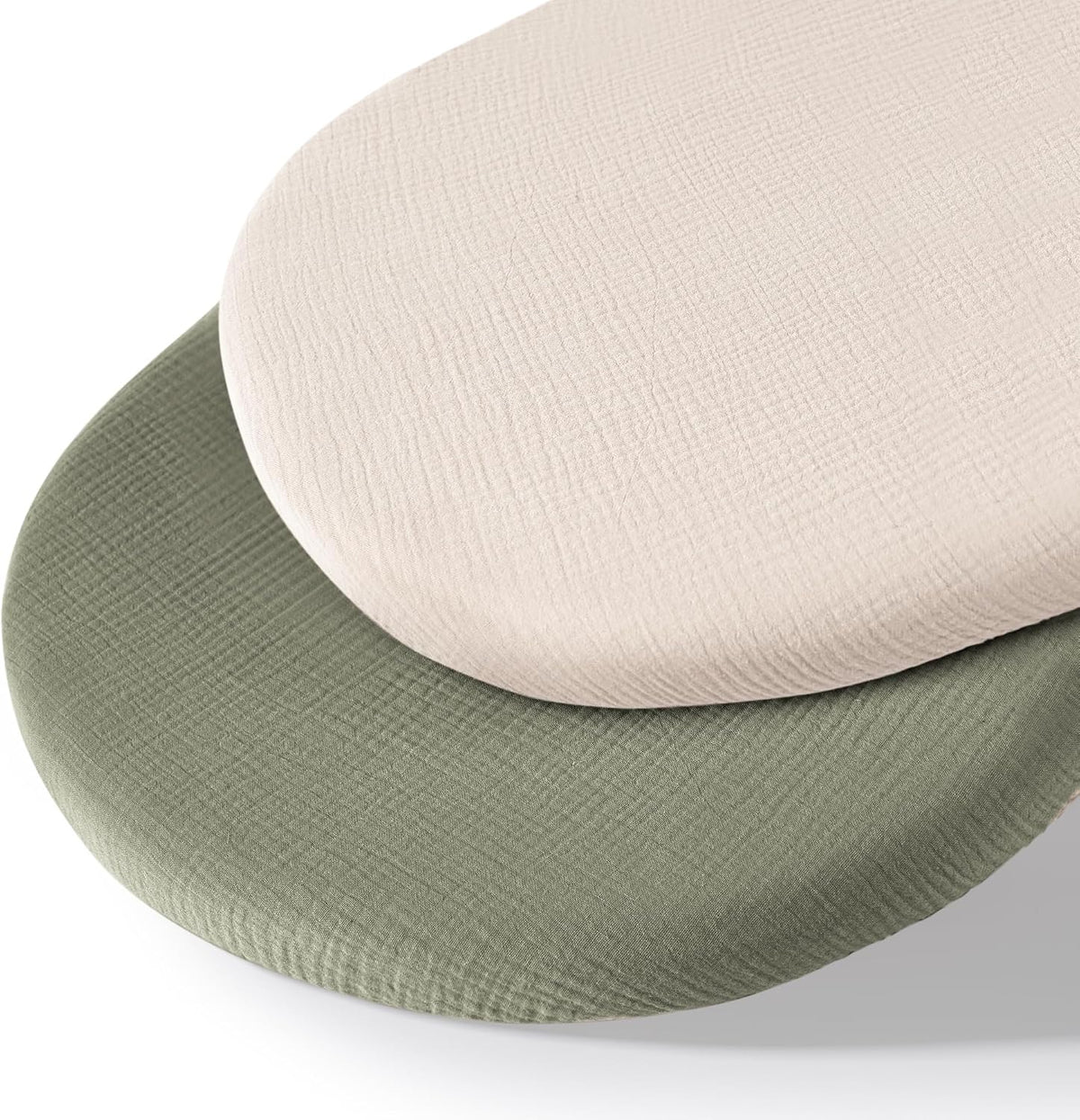 Two oval fitted bassinet sheets stacked at an angle, showing a cream/ivory sheet on top and a sage green sheet beneath. Both feature a crinkled muslin cotton texture on a white background.
