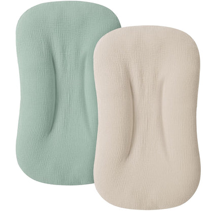Two oval-shaped baby lounger slipcovers with textured muslin fabric, one in sage green and one in ivory/cream, shown overlapping on a white background. Both feature a contoured center indent.