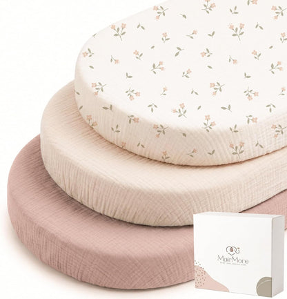 Three stacked oval bassinet fitted sheets by MairMore: top sheet is cream with small pink floral and sage leaf print, middle is solid light cream, bottom is solid dusty rose. Textured muslin fabric visible. White product box shown at lower right.