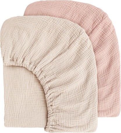 Two muslin fitted crib sheets displayed overlapping: a cream/ivory sheet in front and a dusty pink sheet behind. Both show visible gauze texture and elasticated edges with gathered corners, on a white background.